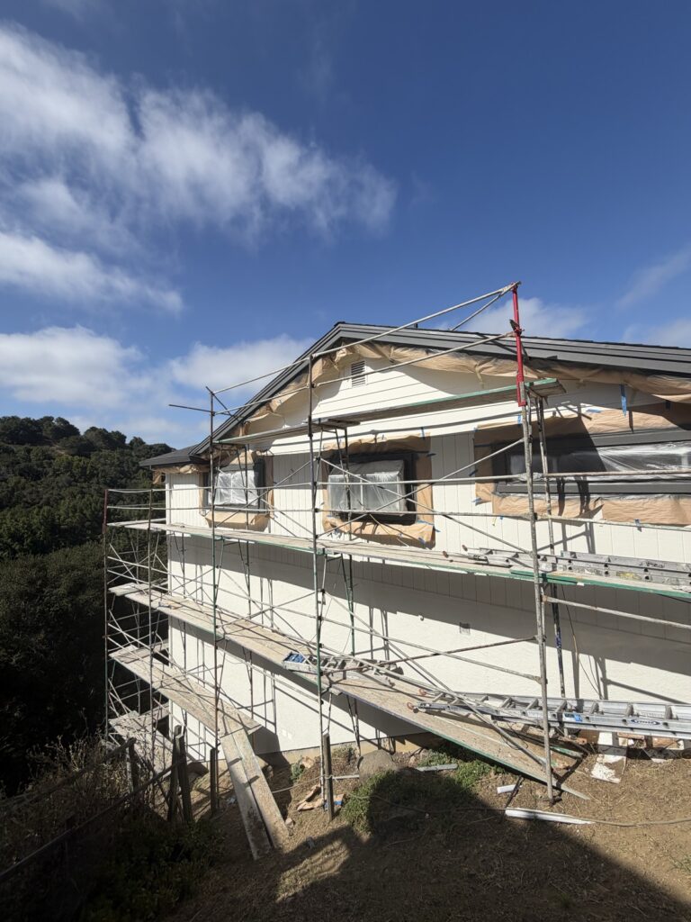 Hillside home exterior painting in progress with scaffolding, cream siding, by Wilmar's Painting